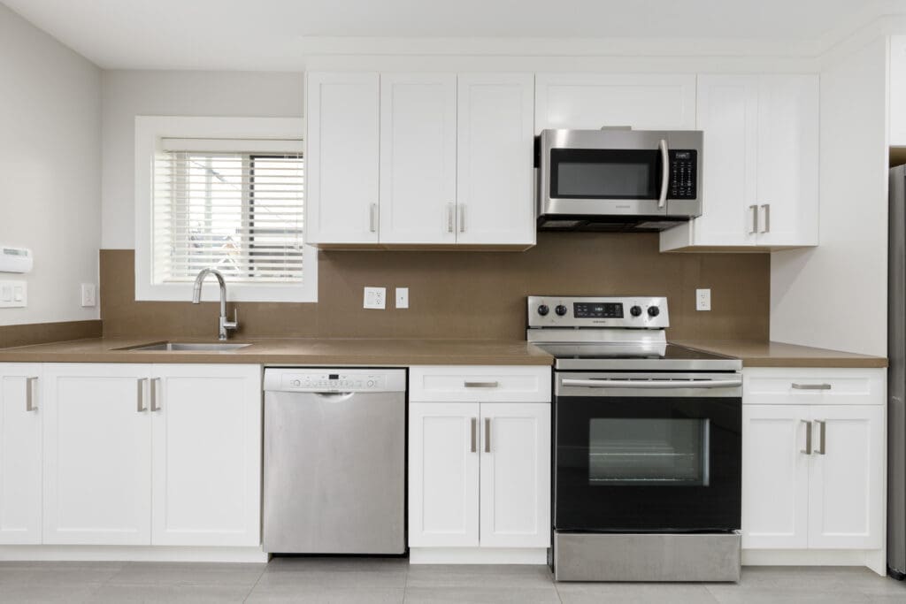 Modern kitchen with white cabinets, stainless steel appliances including a dishwasher, oven, and microwave, a brown backsplash, and a window above the sink.